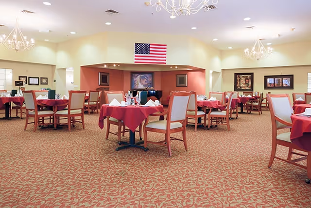 Spacious dining room with multiple tables covered in red tablecloths, chairs, chandeliers, and an American flag on the far wall.
