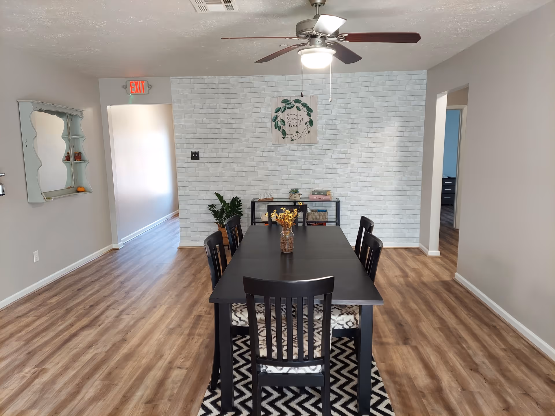 Dining room with a long black table and six chairs on a patterned rug, wood-look floors, ceiling fan, and a white faux-brick accent wall with shelving and wall decor.