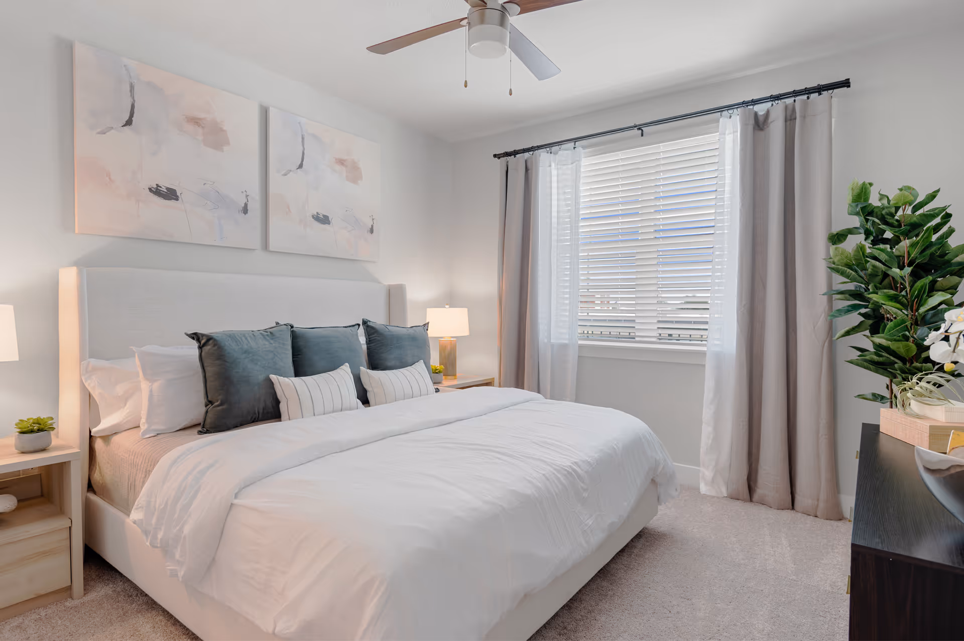 A bright and cozy bedroom with a large bed featuring white bedding and multiple pillows in shades of gray and white. The room has light-colored walls with two abstract paintings above the bed. There are two wooden nightstands with lamps on either side of the bed. A window with white blinds and light gray curtains lets in natural light. A ceiling fan is mounted on the ceiling, and a dark wooden dresser with decorative plants and books is visible on the right side.