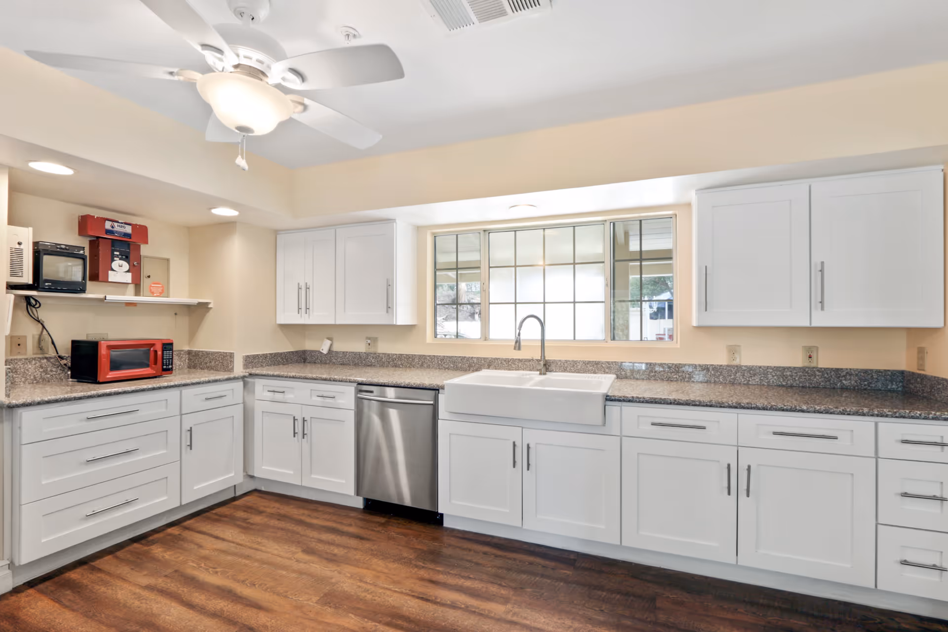 Bright kitchen with white cabinets, granite countertops, a large farmhouse sink under a window, a stainless steel dishwasher, a red microwave, a ceiling fan with light, and wood flooring.