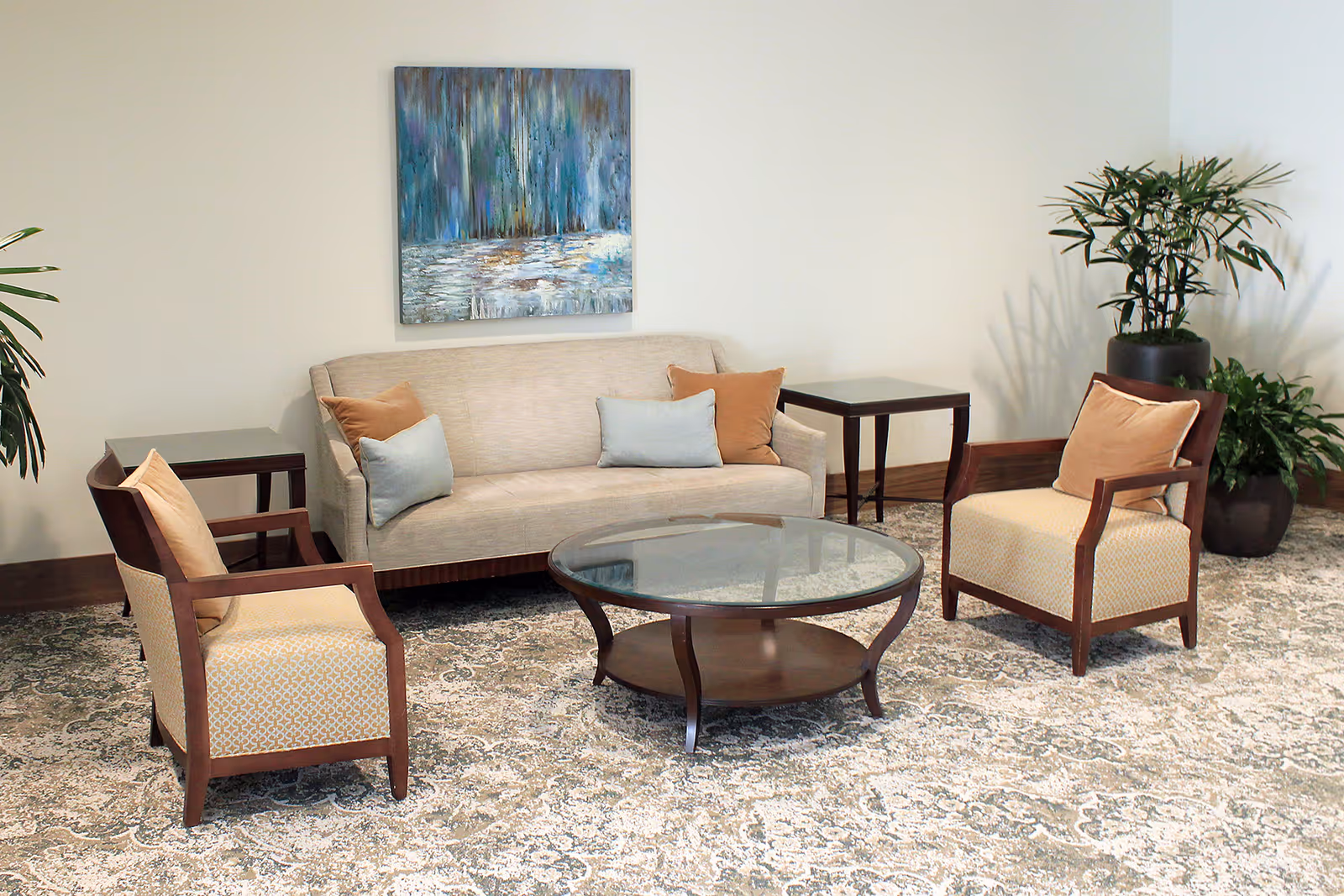 A cozy seating area with a beige sofa adorned with light blue and brown pillows, two matching armchairs with brown wooden frames and beige cushions, a round glass coffee table with a wooden base, two small dark wooden side tables, a large abstract painting on the wall, and two potted plants in the corners.