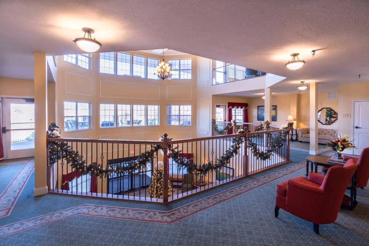 Interior view of a senior living facility's common area decorated for the holidays with garlands and a Christmas tree. The space features large windows, a railing overlooking a lower level, carpeted floors, red armchairs, a coffee table with flowers, and soft lighting fixtures on the ceiling.