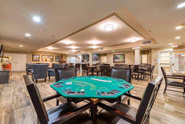 Interior view of a senior living facility common area with a green poker table in the foreground surrounded by chairs. The room has multiple seating areas with tables and chairs, wood-style flooring, and framed artwork on the walls. Ceiling lights illuminate the space, and there is an exit door visible in the background.