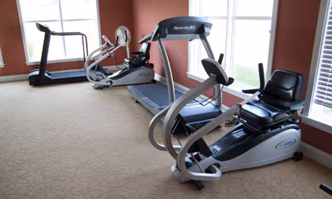A fitness room with exercise equipment including a treadmill, a rowing machine, and a recumbent bike, set against walls painted in a warm reddish color with large windows letting in natural light.