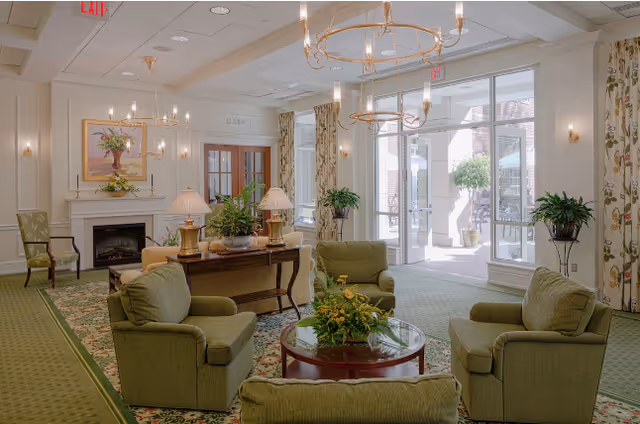 A bright and elegant living room area with green upholstered armchairs arranged around a round glass coffee table with a floral centerpiece. The room features a fireplace with a painting above it, two table lamps on a wooden console table, large windows and glass doors leading to an outdoor patio, floral curtains, and two gold chandeliers hanging from the ceiling.