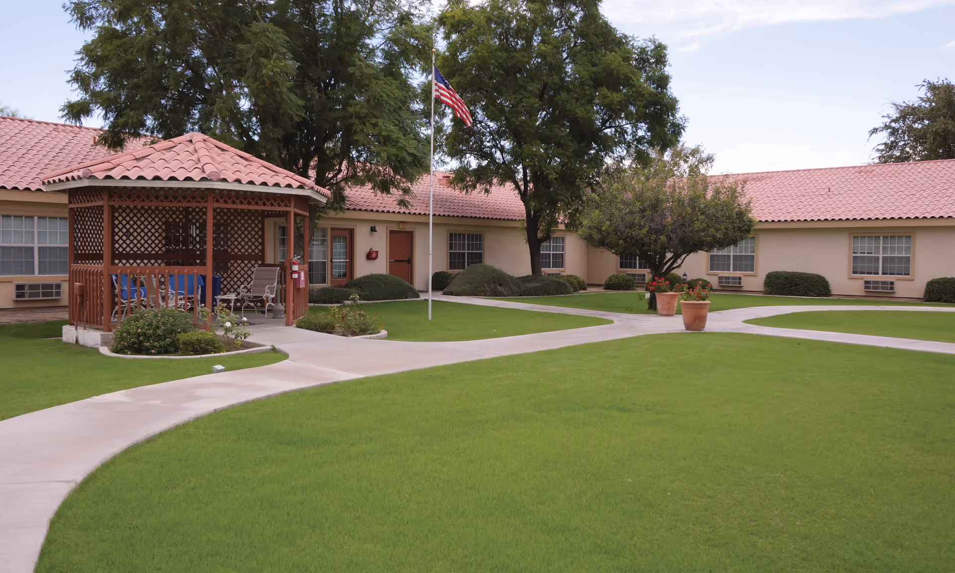 Outdoor courtyard area of a senior living facility with a gazebo, green lawn, paved walkways, trees, potted plants, and a flagpole with an American flag, surrounded by single-story buildings with tiled roofs and windows.