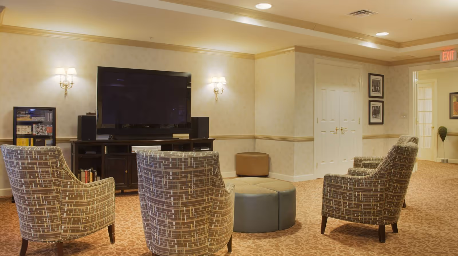 A cozy living room area in a senior living facility with four patterned armchairs arranged around a circular ottoman. A large flat-screen TV is mounted on a dark wooden cabinet against the wall, flanked by two wall sconces with lampshades. The room has beige walls with a chair rail, carpeted floor, and framed pictures on the wall near a double door and an exit sign above a doorway.