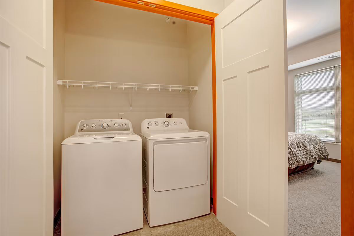 A laundry area with a white washing machine and dryer side by side inside a closet with white sliding doors. A wire shelf is mounted above the machines. To the right, part of a bedroom with a bed and a window with blinds is visible.
