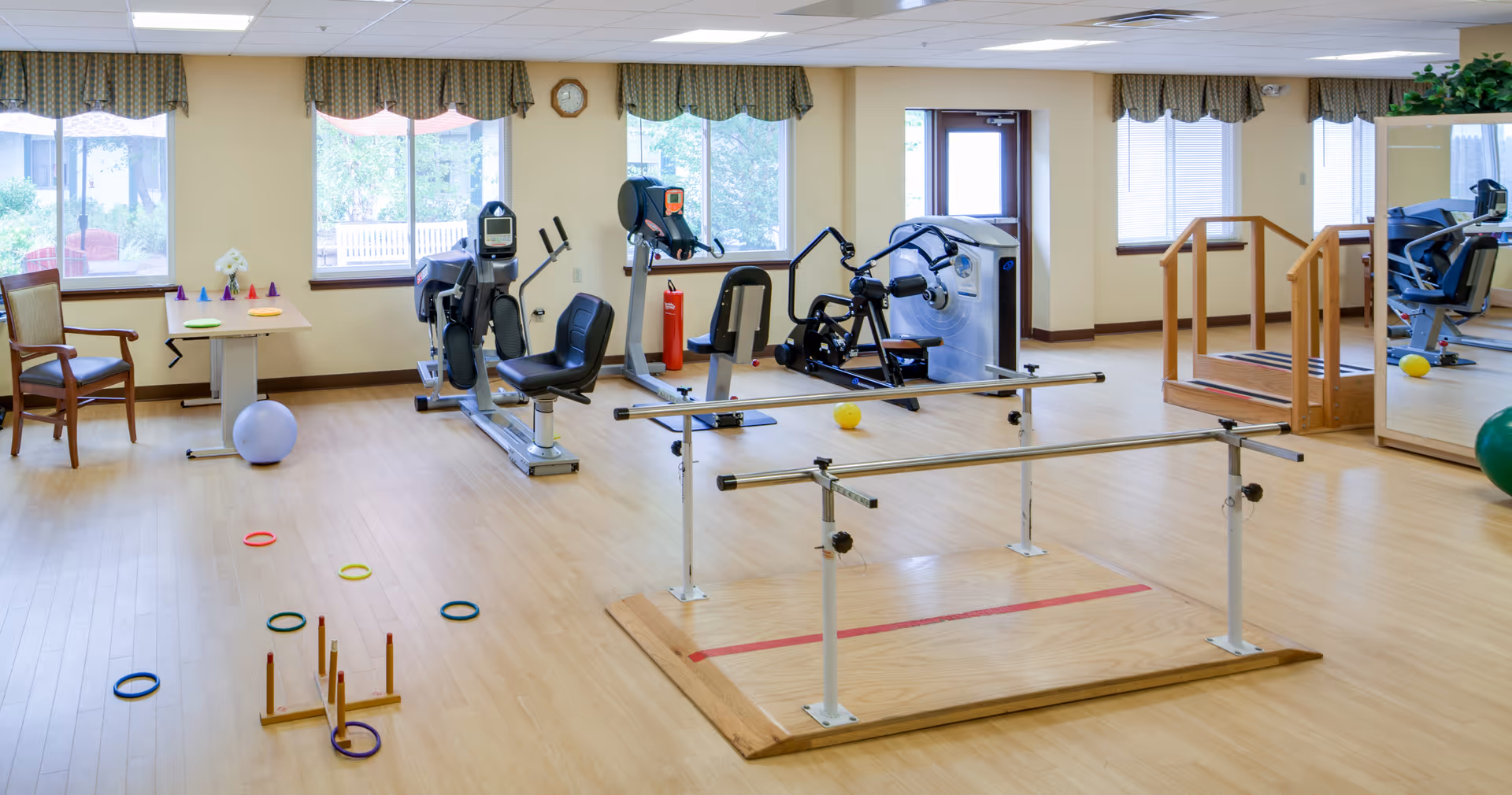 Bright therapy/fitness room with exercise equipment, parallel bars, and mobility aids.