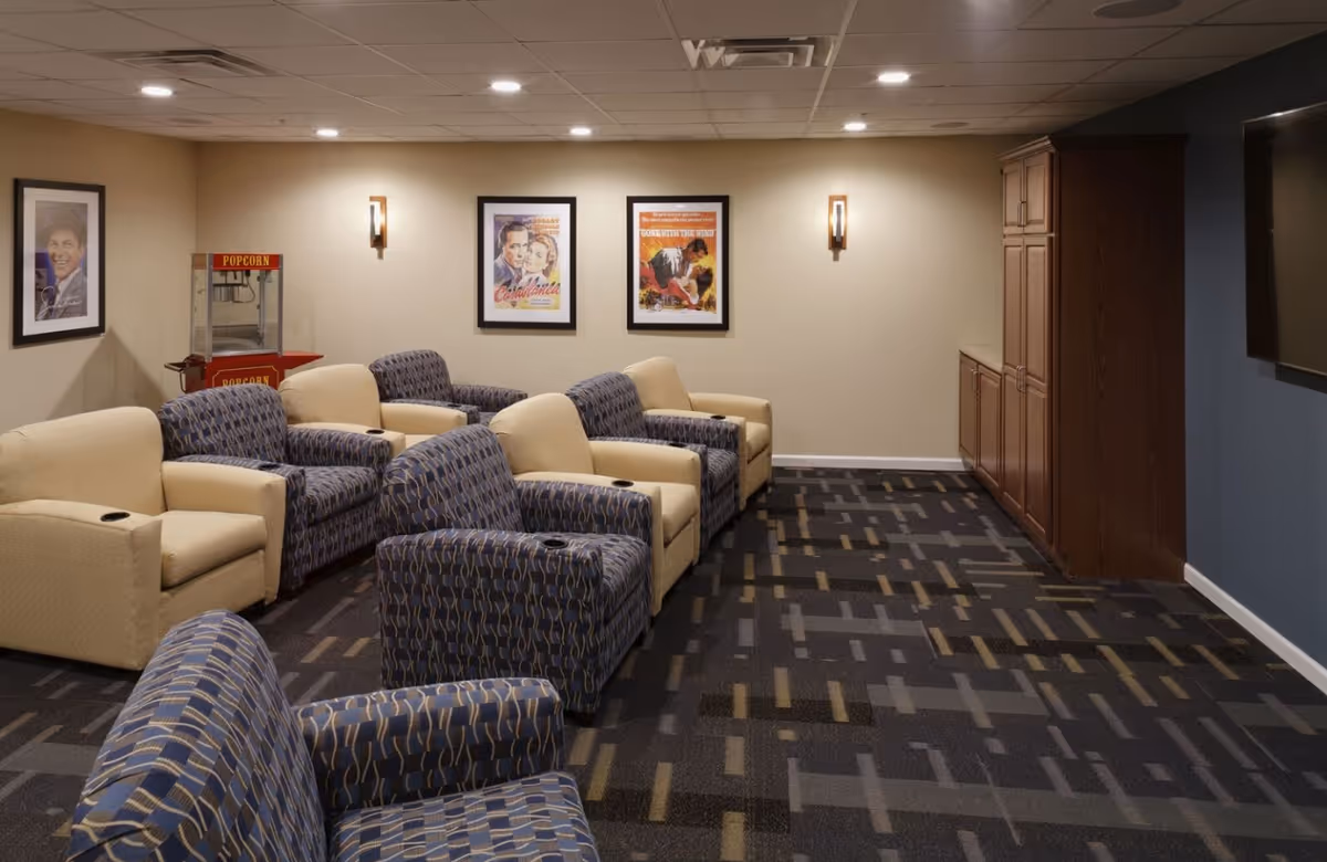 Cozy senior living media room with rows of upholstered recliners, a popcorn machine, framed movie posters, and a wall-mounted TV.