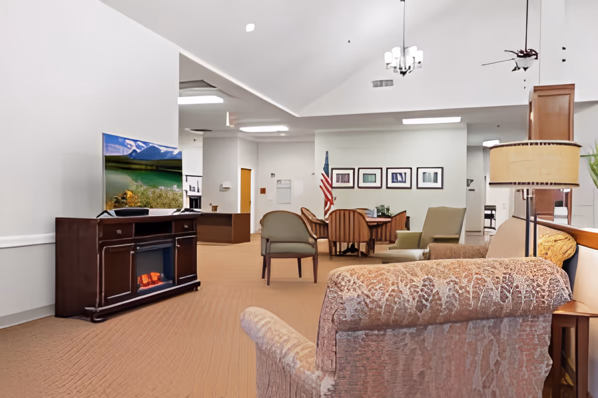 Spacious senior living common lounge with upholstered chairs, a TV on a console, a dining table in the background, and soft overhead lighting.