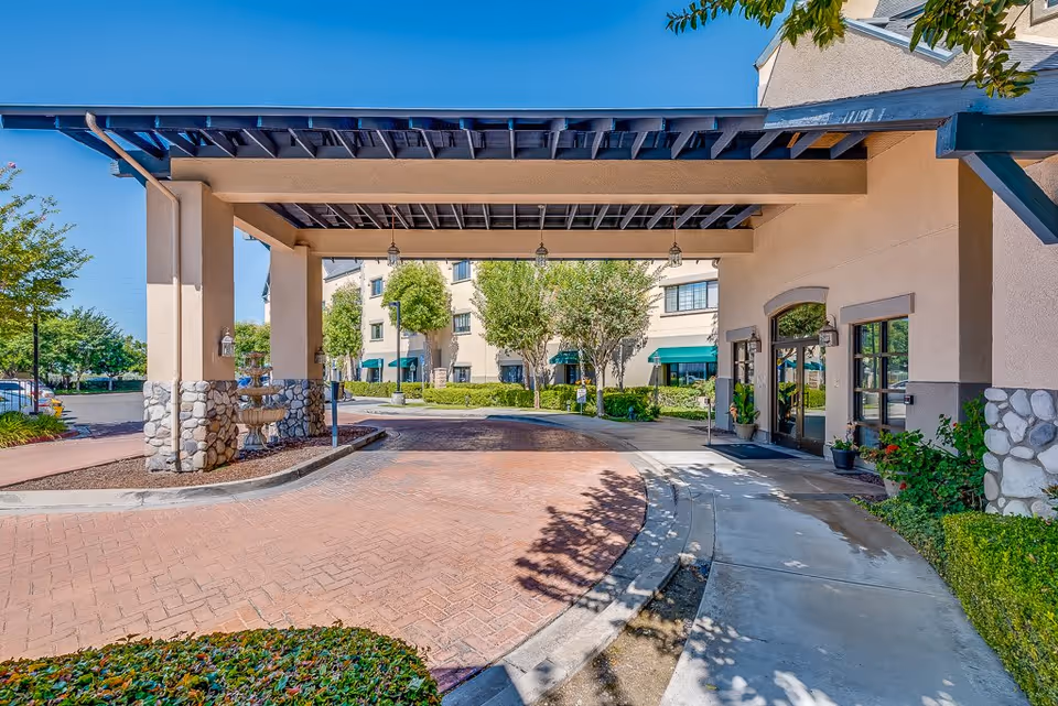 Covered driveway entrance to a senior living facility with stone pillars and hanging lanterns. The building has beige walls with green awnings over the windows and doors. There are trees and shrubs around the entrance and a clear blue sky above.