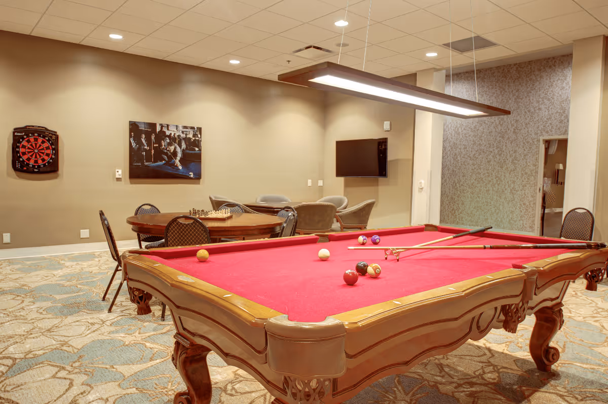 A recreational room with a red felt pool table in the foreground, pool cues resting on it, and several billiard balls scattered on the table. In the background, there is a round table with chairs and a chessboard, a dartboard mounted on the wall, a flat-screen TV, and additional seating. The room has beige walls, a patterned carpet, and ceiling lights.