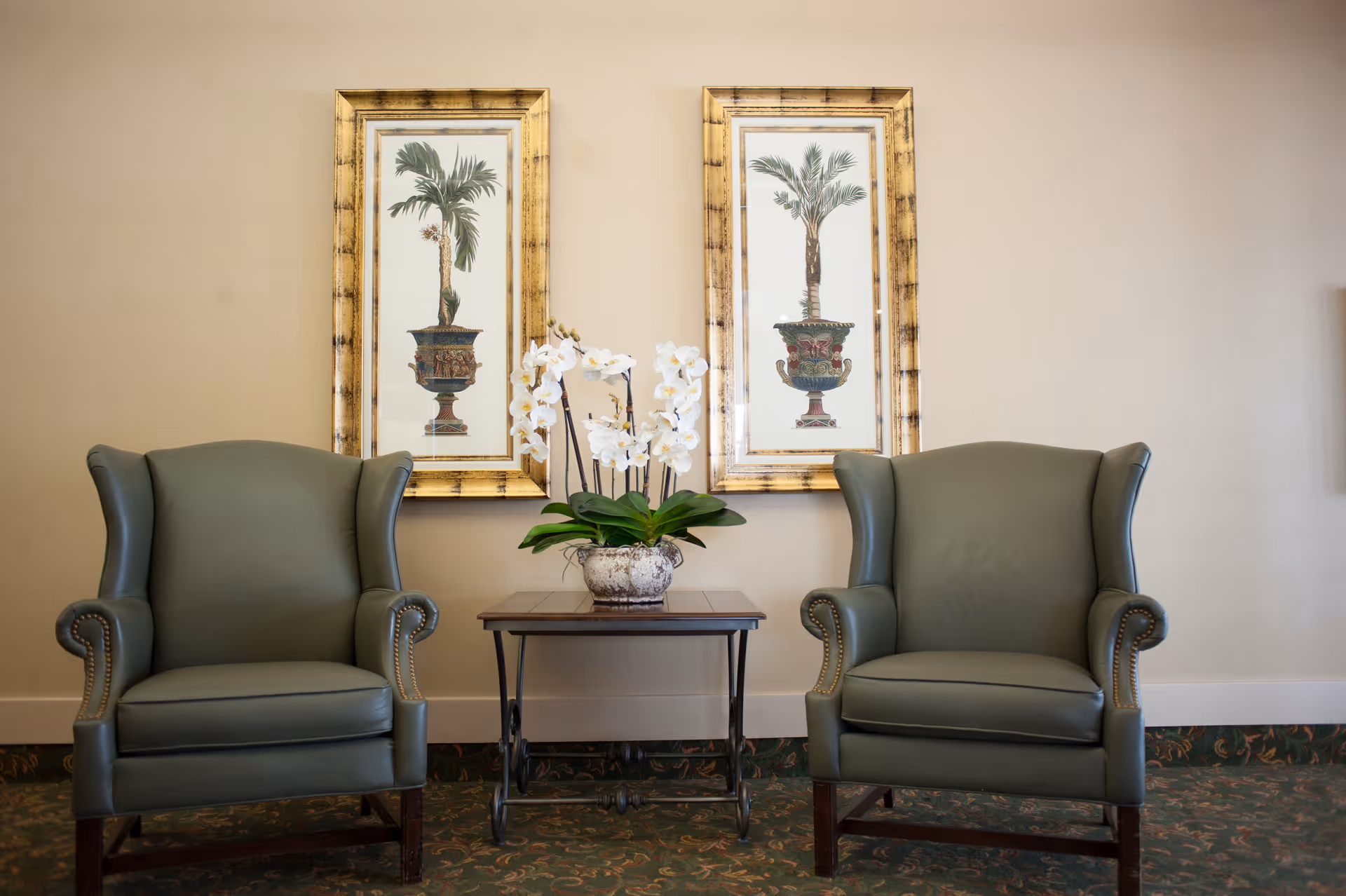 Two green upholstered armchairs with wooden legs are positioned on either side of a small wooden table holding a white orchid plant in a decorative pot. Behind the chairs, two framed botanical prints of potted palm plants hang on a beige wall. The floor is covered with a patterned carpet.