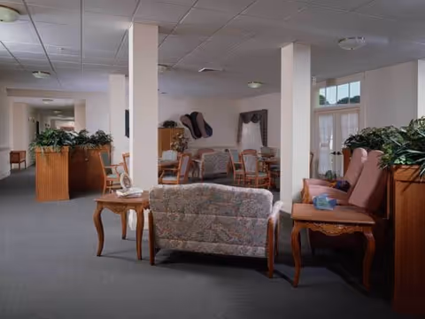 A spacious senior living facility lounge with upholstered sofas and armchairs, wooden side tables, potted plants, and supportive columns.