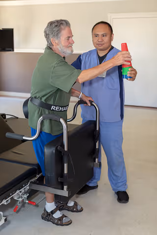 An elderly man wearing a green shirt and blue pants is using a walker labeled 'REHAB' while a healthcare professional in blue scrubs assists him by holding colorful stacking cups in a rehabilitation or therapy room.