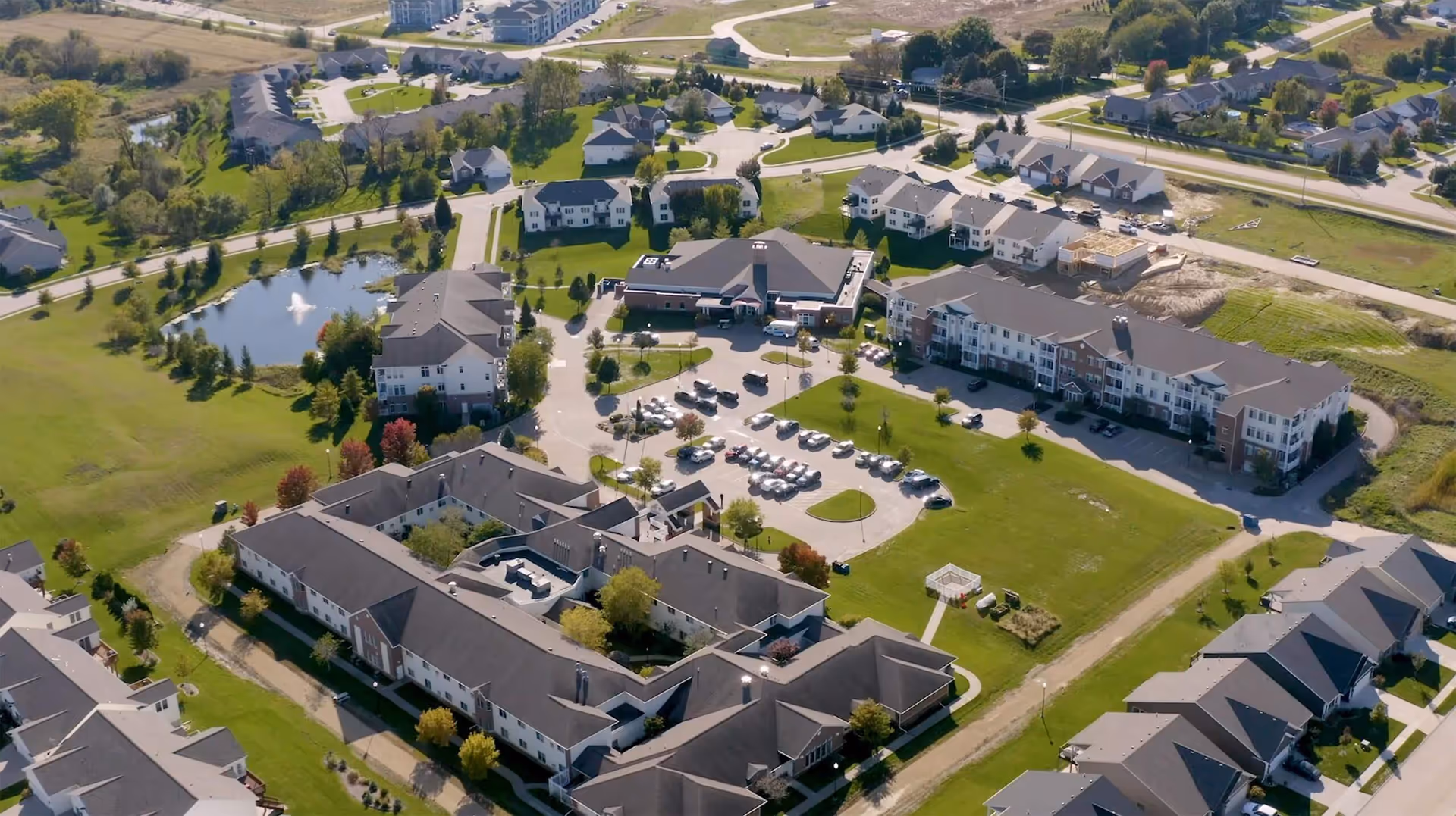 Aerial view of The Fountains senior living facility showing multiple buildings, parking lots with cars, green lawns, trees, a small pond with a fountain, and surrounding residential neighborhood.