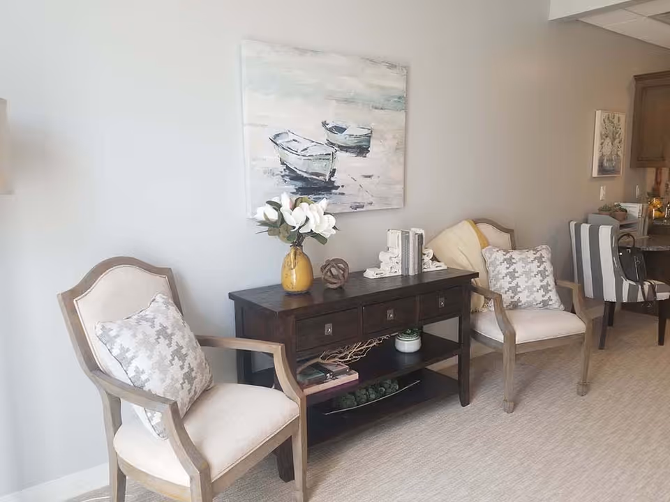 A cozy interior space featuring two beige cushioned armchairs with patterned pillows, a dark wooden console table with decorative items including a yellow vase with white flowers, books, and a small plant. Above the table hangs a painting of two boats on water. In the background, part of a kitchen area with striped chairs is visible.