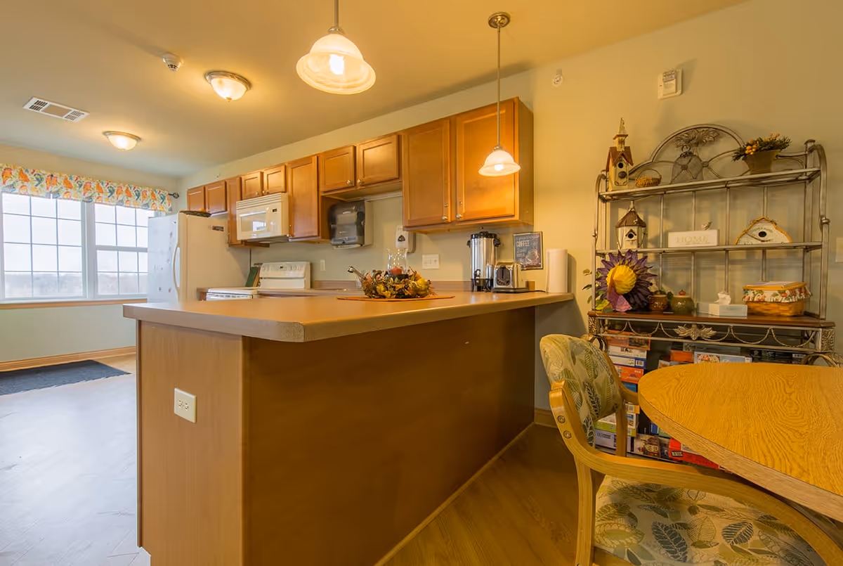 A bright kitchen area with wooden cabinets, a white refrigerator, microwave, and stove. A kitchen island with a decorative centerpiece is in the foreground. To the right, there is a metal shelving unit with decorative items and board games, and a round wooden table with a cushioned chair featuring a leaf pattern. Large windows with floral valances allow natural light into the room.