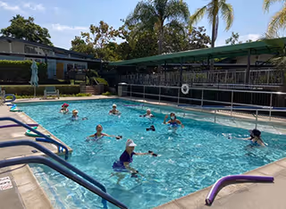 Outdoor swimming pool with several elderly people participating in a water exercise class. The pool is surrounded by lounge chairs and shaded seating areas. Palm trees and other greenery are visible in the background under a clear sky.
