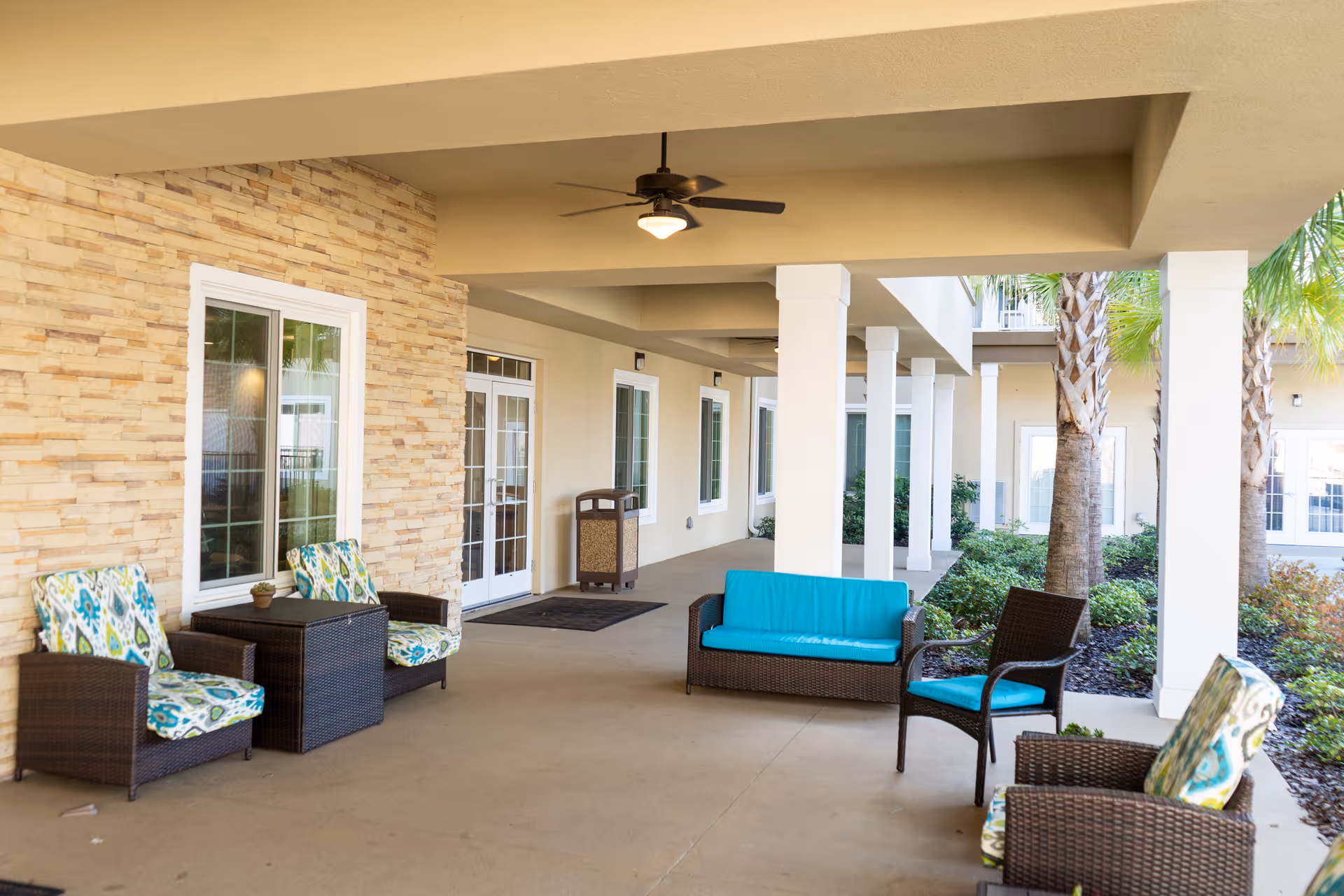 Covered outdoor patio area with wicker furniture including chairs with patterned cushions and a loveseat with a blue cushion, ceiling fans, and a stone accent wall with windows and doors leading inside. Palm trees and greenery are visible along the edge of the patio.