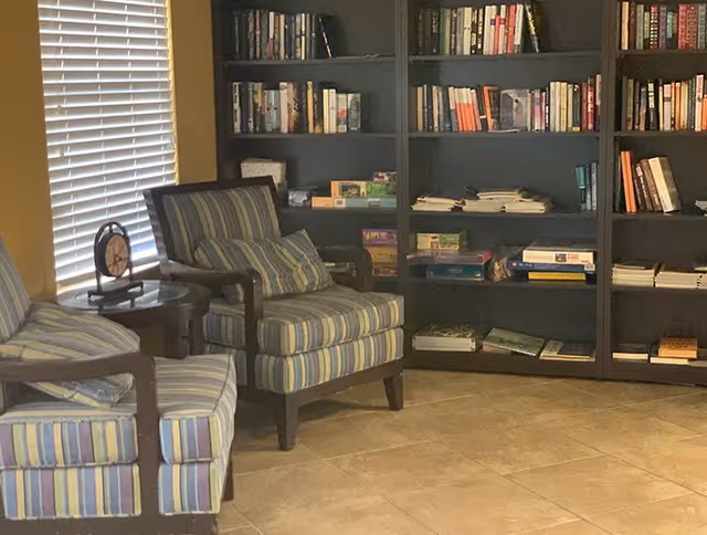 A cozy sitting area with two striped armchairs and a small round wooden table between them, placed next to a window with closed blinds. Behind the chairs is a large dark wooden bookshelf filled with books and board games.