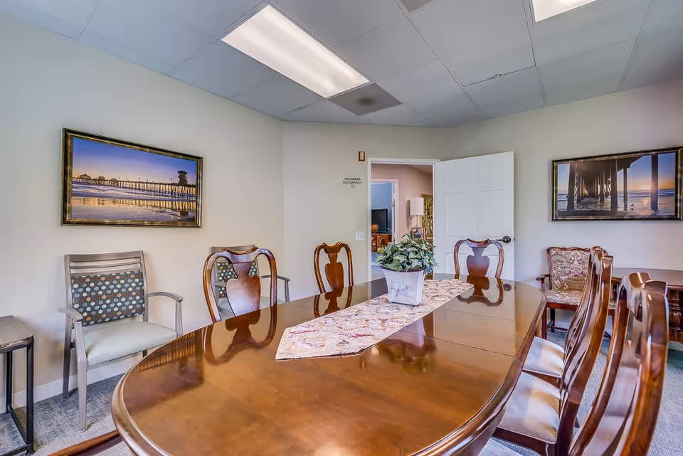 A well-lit dining room with a large polished wooden table surrounded by wooden chairs with cushioned seats. A decorative runner and a potted plant are placed on the table. The walls are adorned with framed pictures of piers over water. An open door leads to another room with a television and additional furniture.