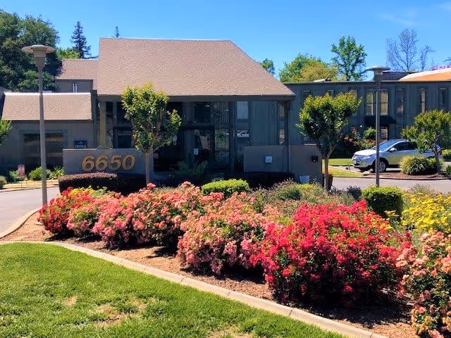 Exterior view of a senior living facility building with the address number 6650 displayed on a sign surrounded by colorful flowering bushes and green grass. The building has large windows and a sloped roof, with a parking area and cars visible in the background under a clear blue sky.