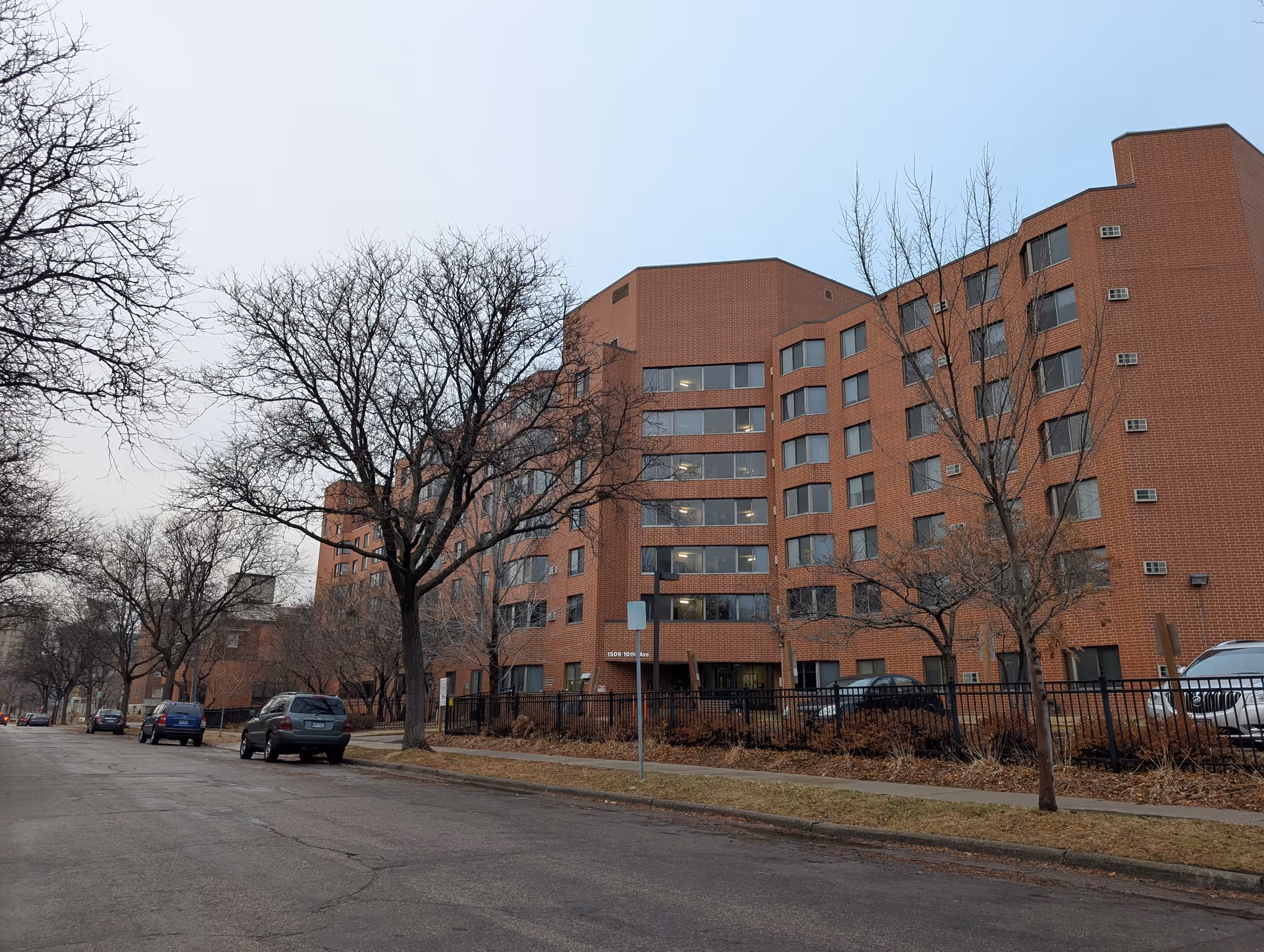 Exterior view of a multi-story brick apartment building with several windows and a fenced area in front. Leafless trees line the sidewalk along a quiet street with parked cars.