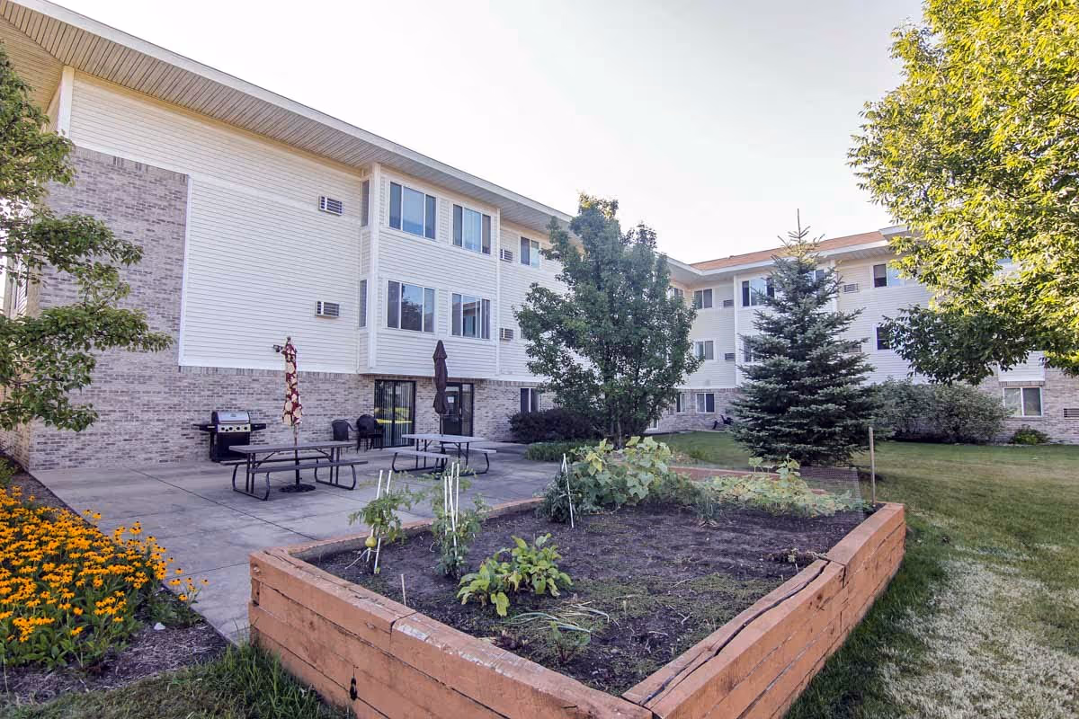 Outdoor view of Glenwood Senior Apartments showing a raised garden bed with plants, a concrete patio with picnic tables and umbrellas, a barbecue grill, and a three-story building with multiple windows in the background surrounded by trees and grass.