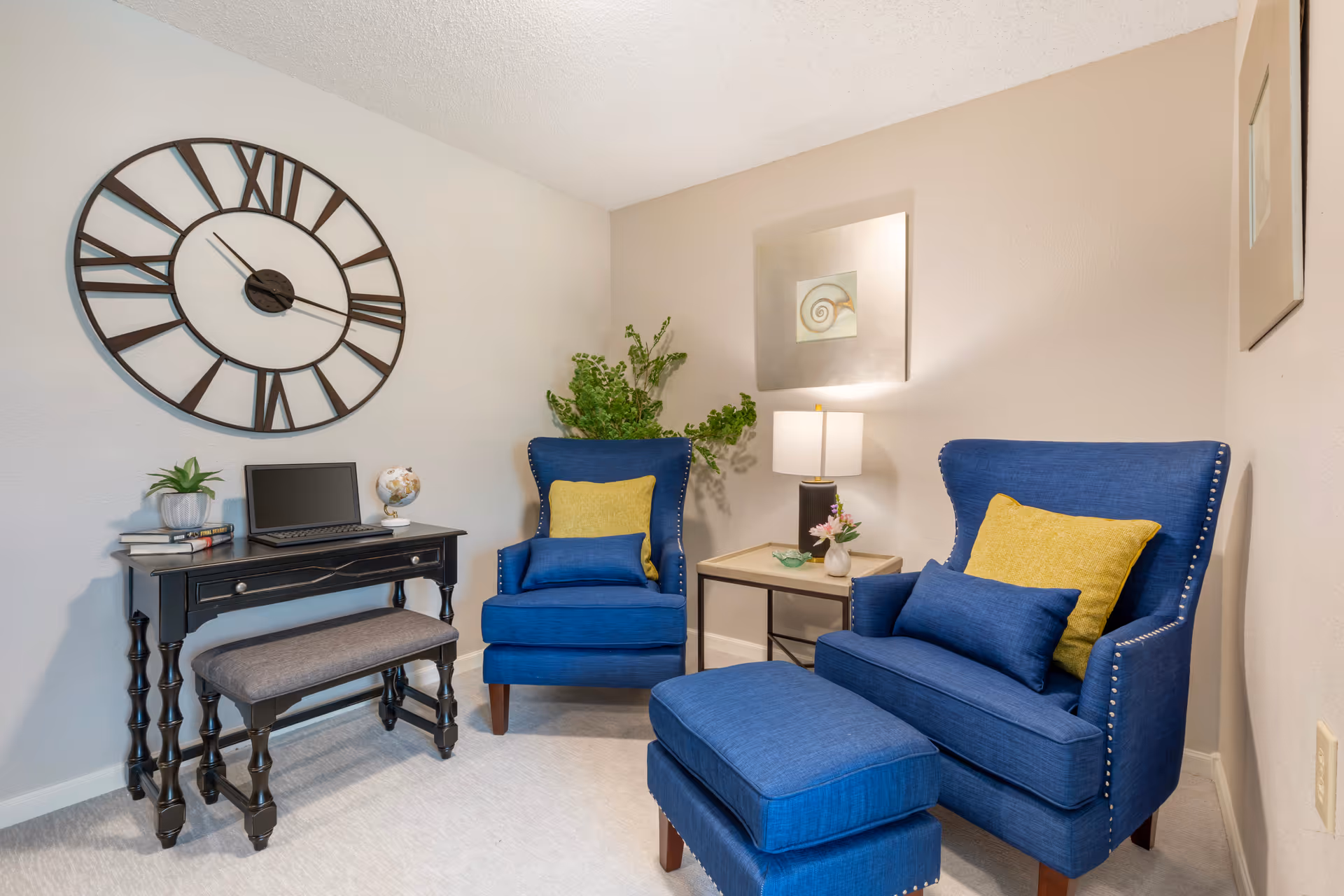 Cozy sitting area with two blue armchairs and an ottoman, a small side table and a desk beneath a large wall clock.