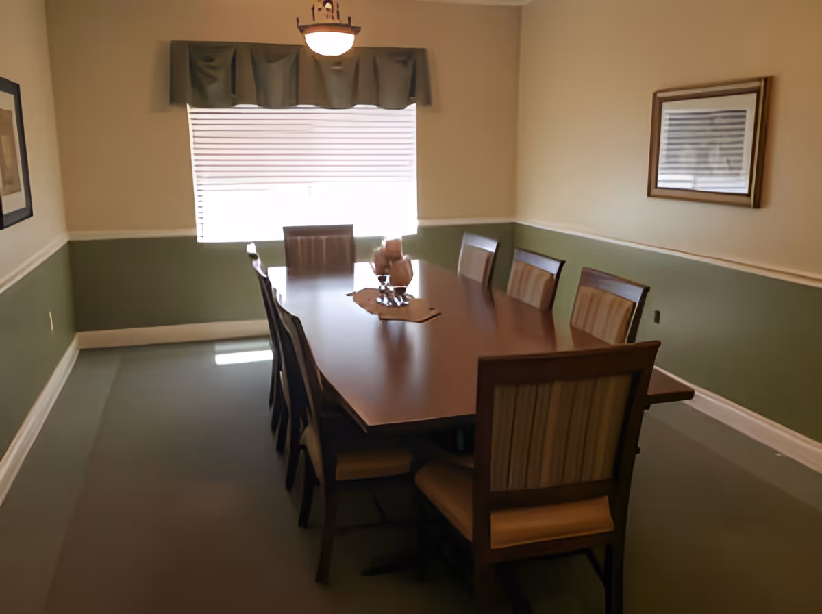 A dining room with a long wooden table surrounded by eight chairs with striped upholstery. The walls are painted beige on the upper half and green on the lower half, separated by a white chair rail. A window with closed blinds and a green valance is at the far end of the room. There is a framed picture on the right wall and a ceiling light fixture above the table.