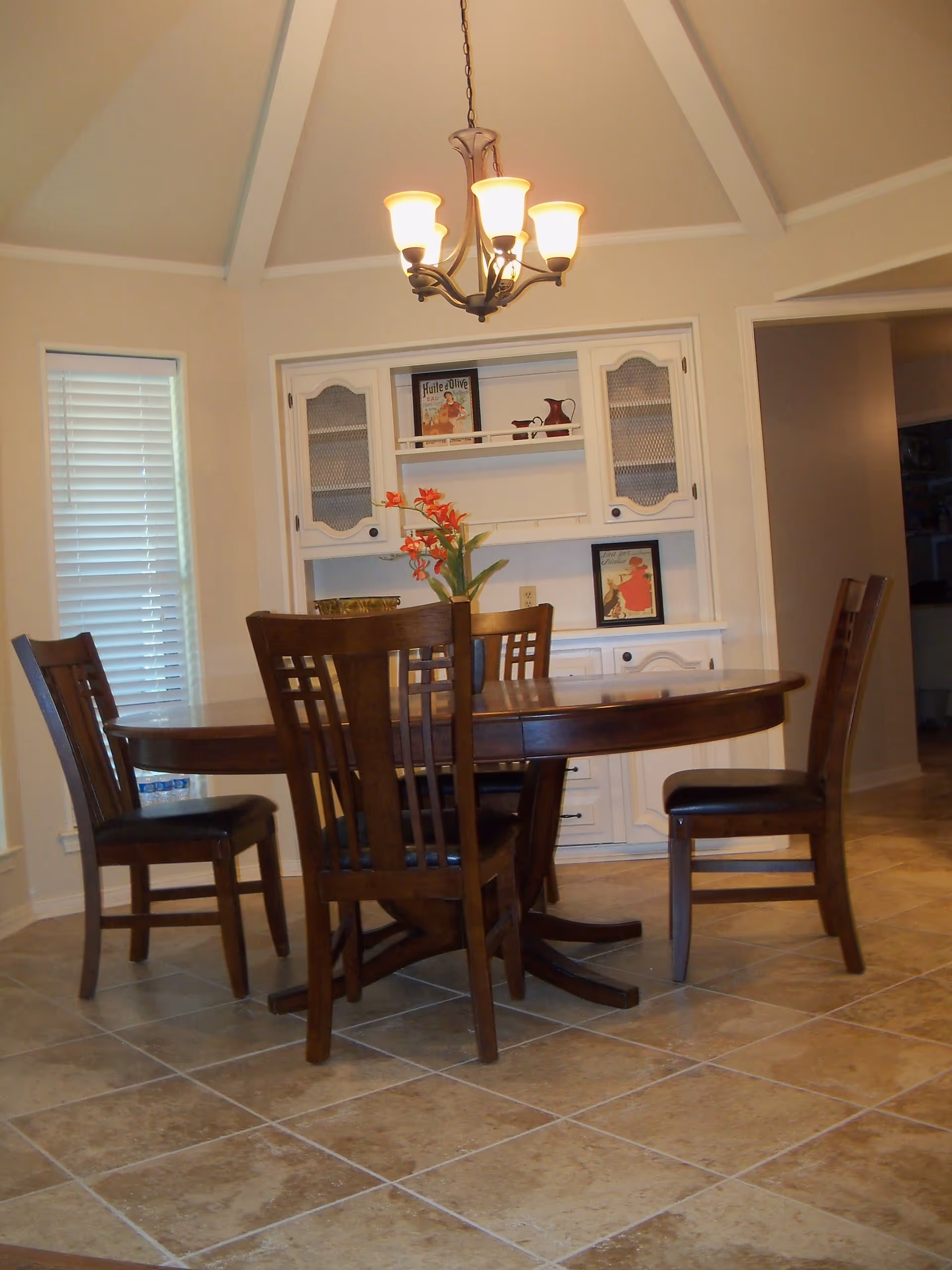 A dining room with a round wooden table surrounded by four wooden chairs with dark cushions. A vase with red flowers is placed on the table. The room has beige tiled flooring and cream-colored walls. A chandelier with five lights hangs from the ceiling. In the background, there is a white built-in cabinet with glass-paneled doors and decorative items on the shelves. A window with closed blinds is visible on the left side.