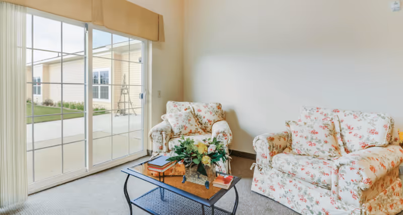 Sunlit living area with two floral upholstered armchairs, a glass-top coffee table with books and a floral arrangement, and a sliding glass door to the outside.