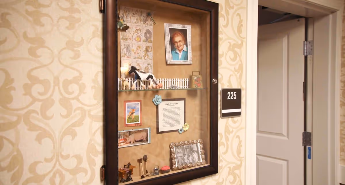 A hallway wall display case with a framed photo of an elderly woman, small animal figurines, and various pictures and notes. The wall has a beige patterned wallpaper, and a door labeled 225 is partially open nearby.