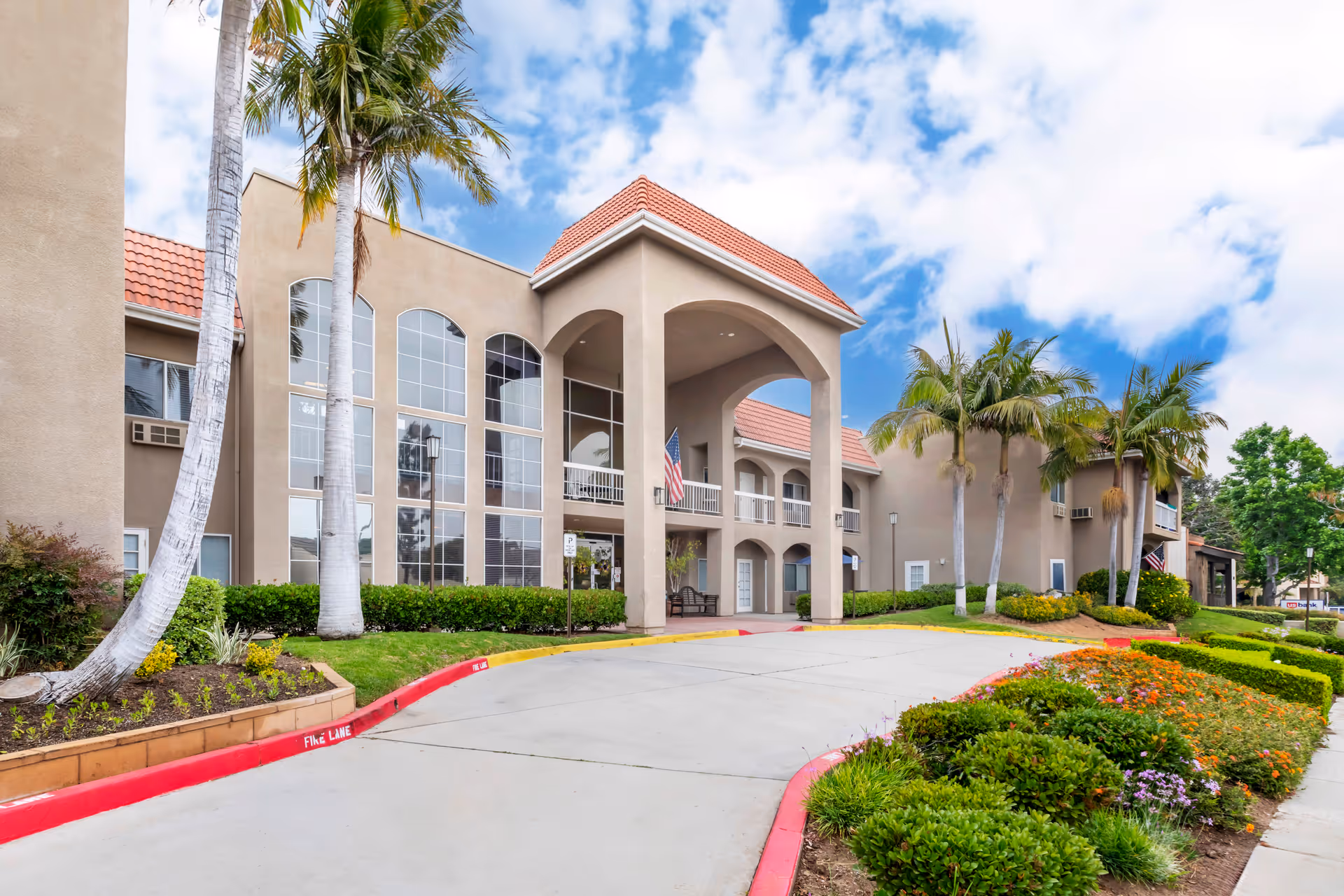 Exterior view of Brookdale Brea senior living facility showing a beige building with large arched windows, a covered entrance with columns, palm trees, and well-maintained landscaping under a partly cloudy sky.