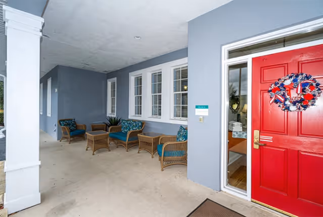 Covered front entry porch with a bright red door, decorative wreath, wicker seating with blue cushions, and a row of windows along a gray exterior wall.