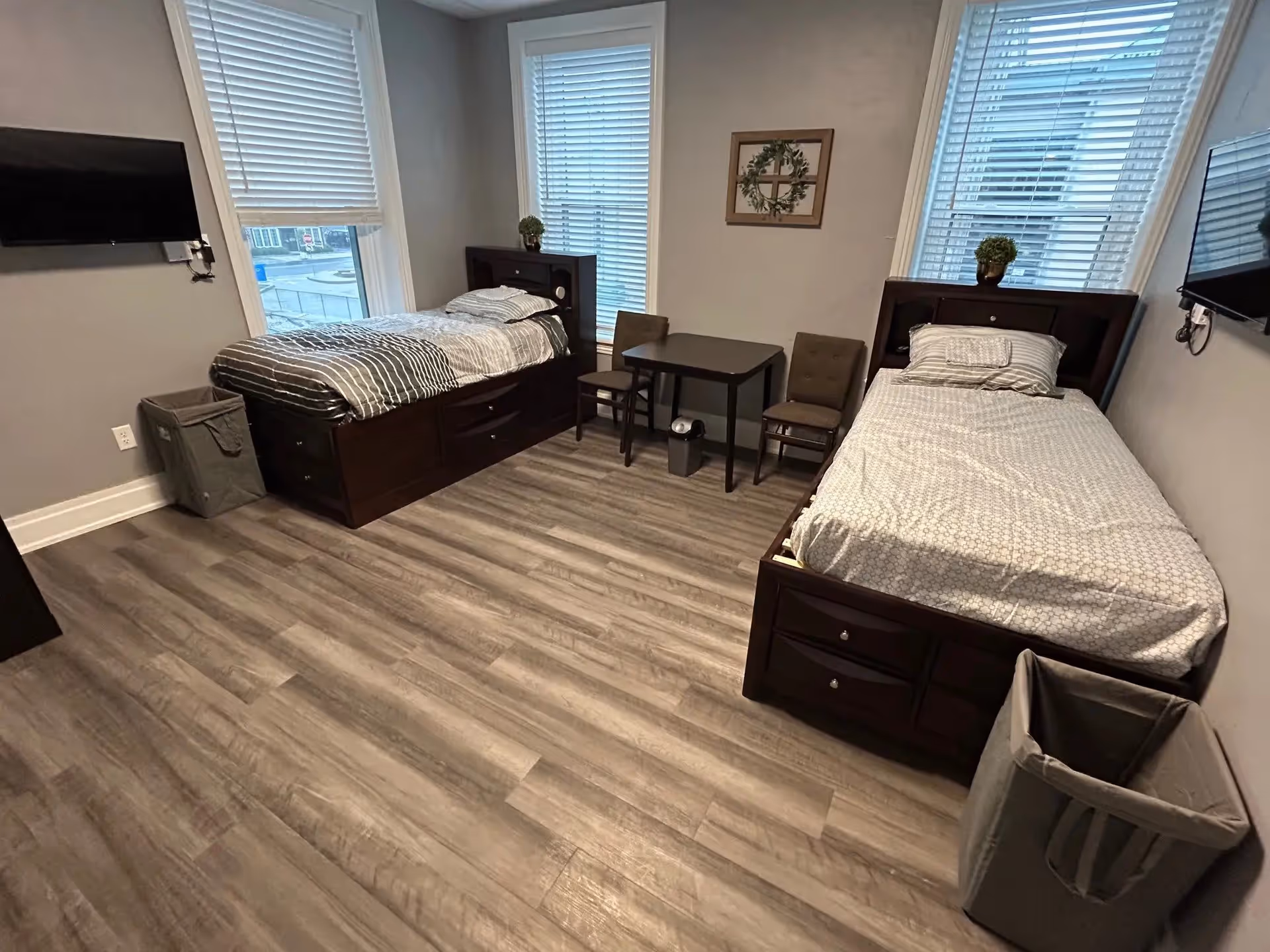 A bedroom with two single beds placed against opposite walls, each with a dark wooden frame and storage drawers underneath. Between the beds is a small dark wooden table with two chairs. The room has light gray walls, two large windows with white blinds, a wall-mounted TV on each side, and a small decorative plant on each headboard. There is a small trash bin near each bed and a framed wall decoration with a cross and greenery above the table.