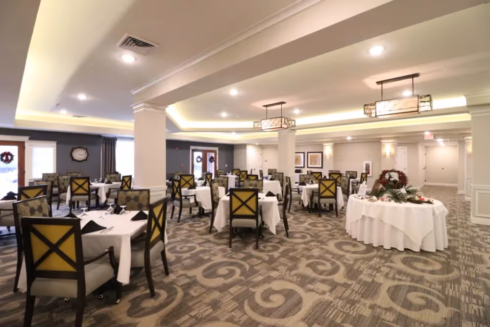 A spacious dining room with multiple tables covered in white tablecloths, each set with black napkins and glassware. The room features patterned carpet flooring, cushioned chairs with wooden frames, and soft ceiling lighting with decorative hanging fixtures. There is a round table with a white tablecloth and holiday wreath decorations near the right side of the room.