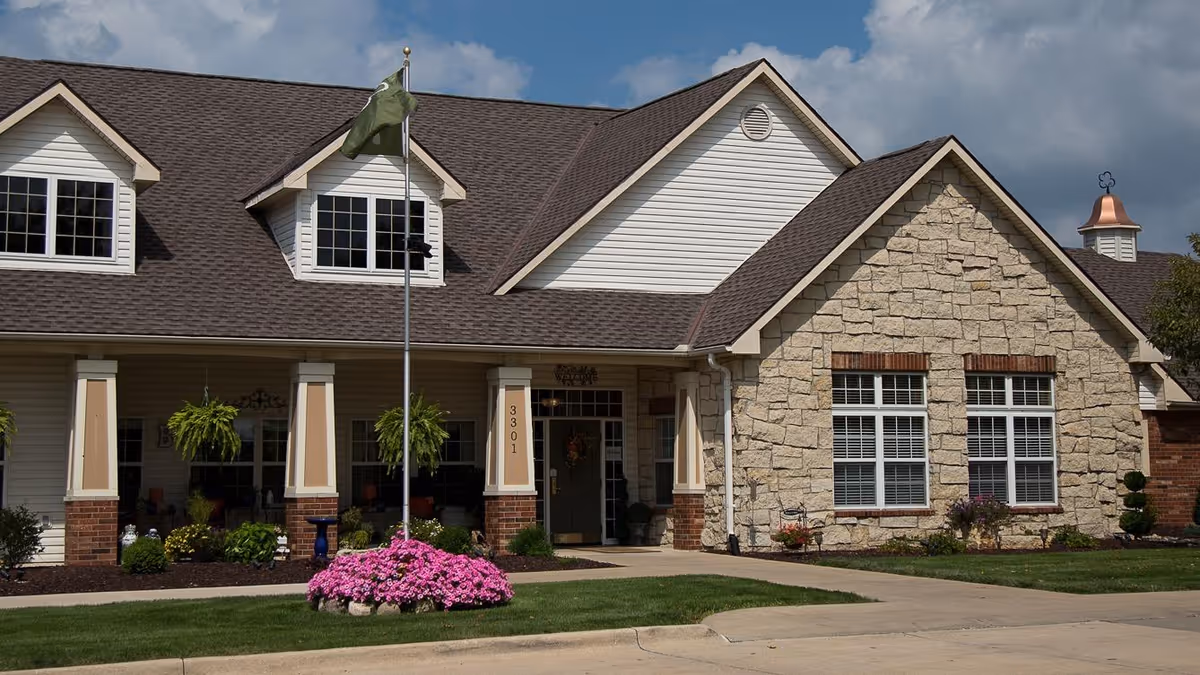 Front exterior of a single-story senior living building with a covered entrance, flagpole, and landscaped flower beds.