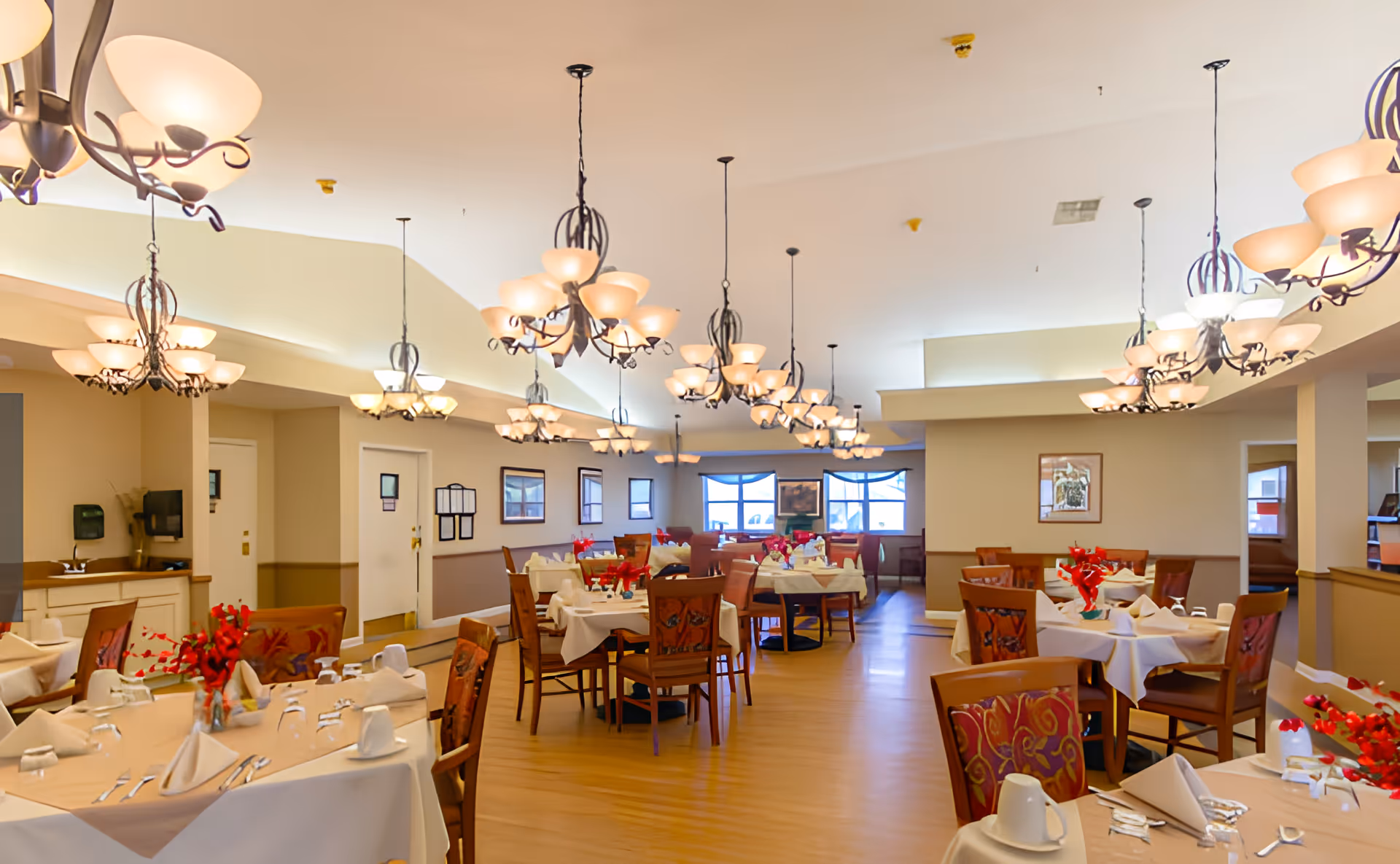 Spacious dining room with multiple set tables, hanging chandeliers, and red floral centerpieces.