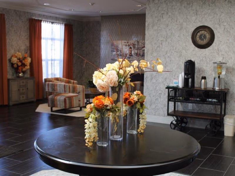 A cozy interior common area with a round black table in the foreground holding three glass vases filled with artificial flowers in shades of orange, cream, and yellow. In the background, there are two striped armchairs near a window with orange curtains, a small cabinet with a flower arrangement, a wall clock, and a refreshment station with a coffee maker and water dispenser on a black cart.
