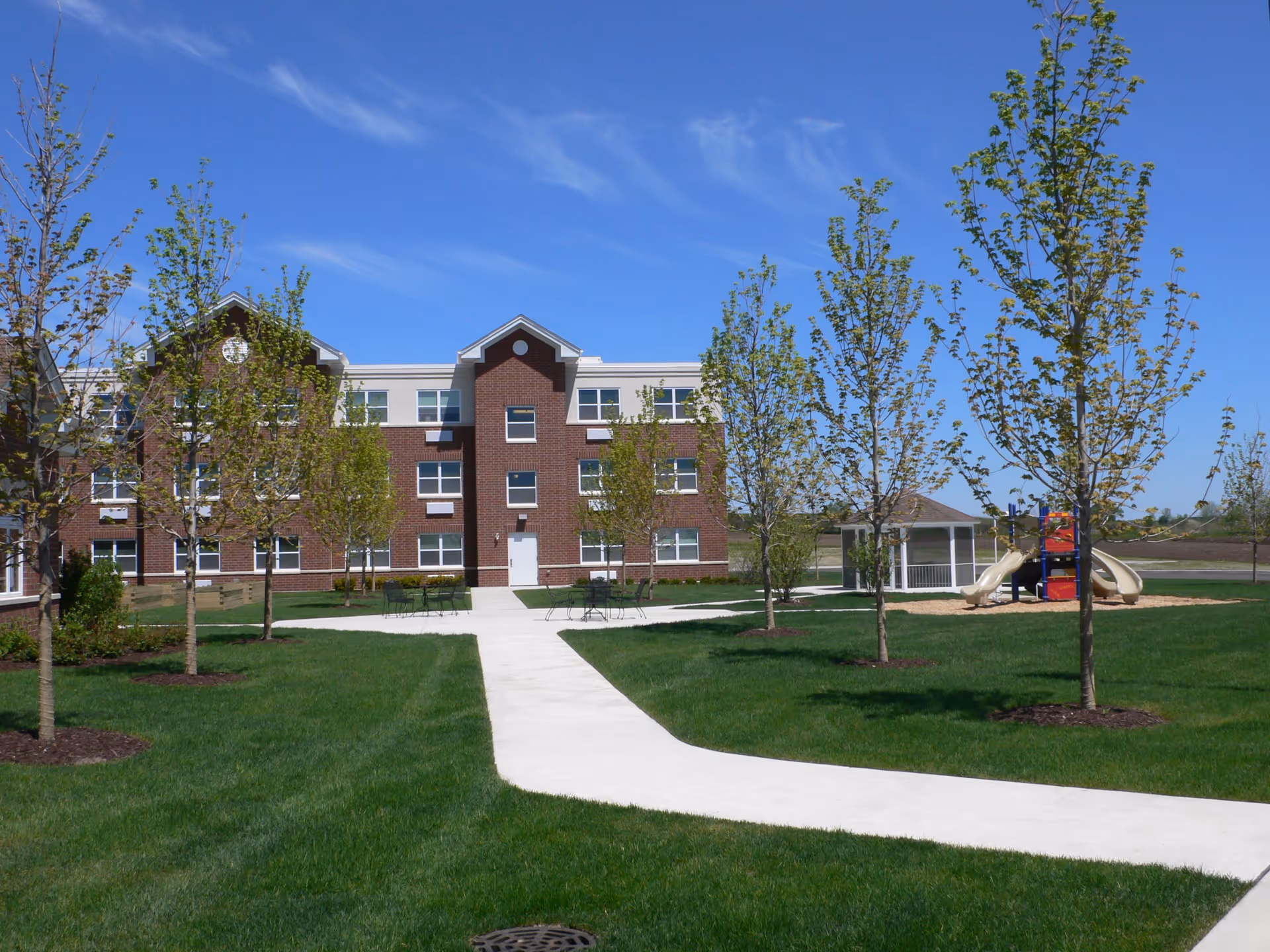 Outdoor view of a senior living facility with a three-story brick and beige building in the background. The foreground features a well-maintained lawn with young trees planted in mulch beds, a concrete walkway, several metal tables and chairs, a gazebo, and a playground with slides. The sky is clear and blue.