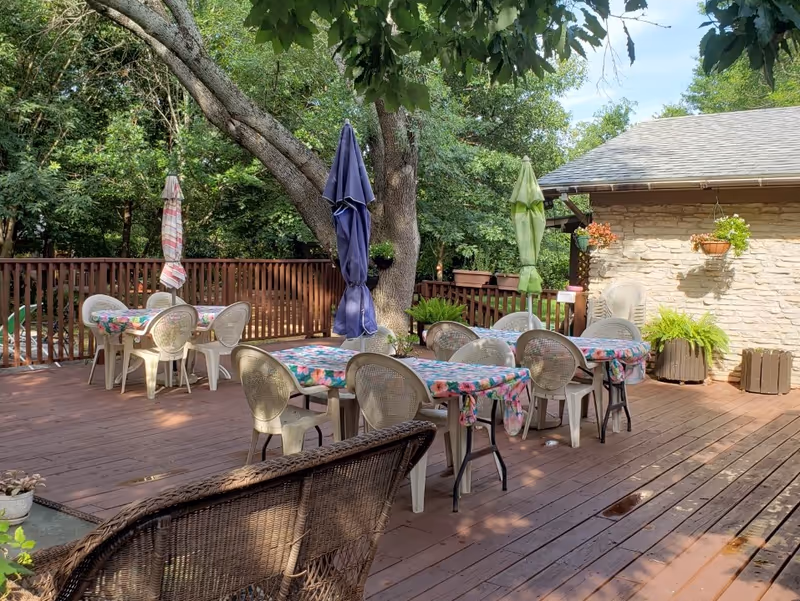 Wooden outdoor deck with multiple tables and chairs covered in floral tablecloths, closed umbrellas, hanging plants, and surrounding trees.