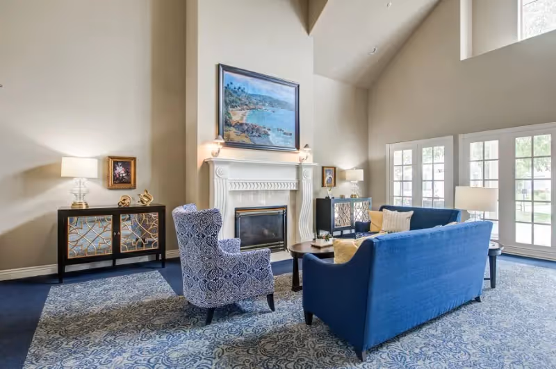 Bright, spacious living room with blue sofas and patterned armchair arranged around a fireplace beneath a large painting and tall windows.