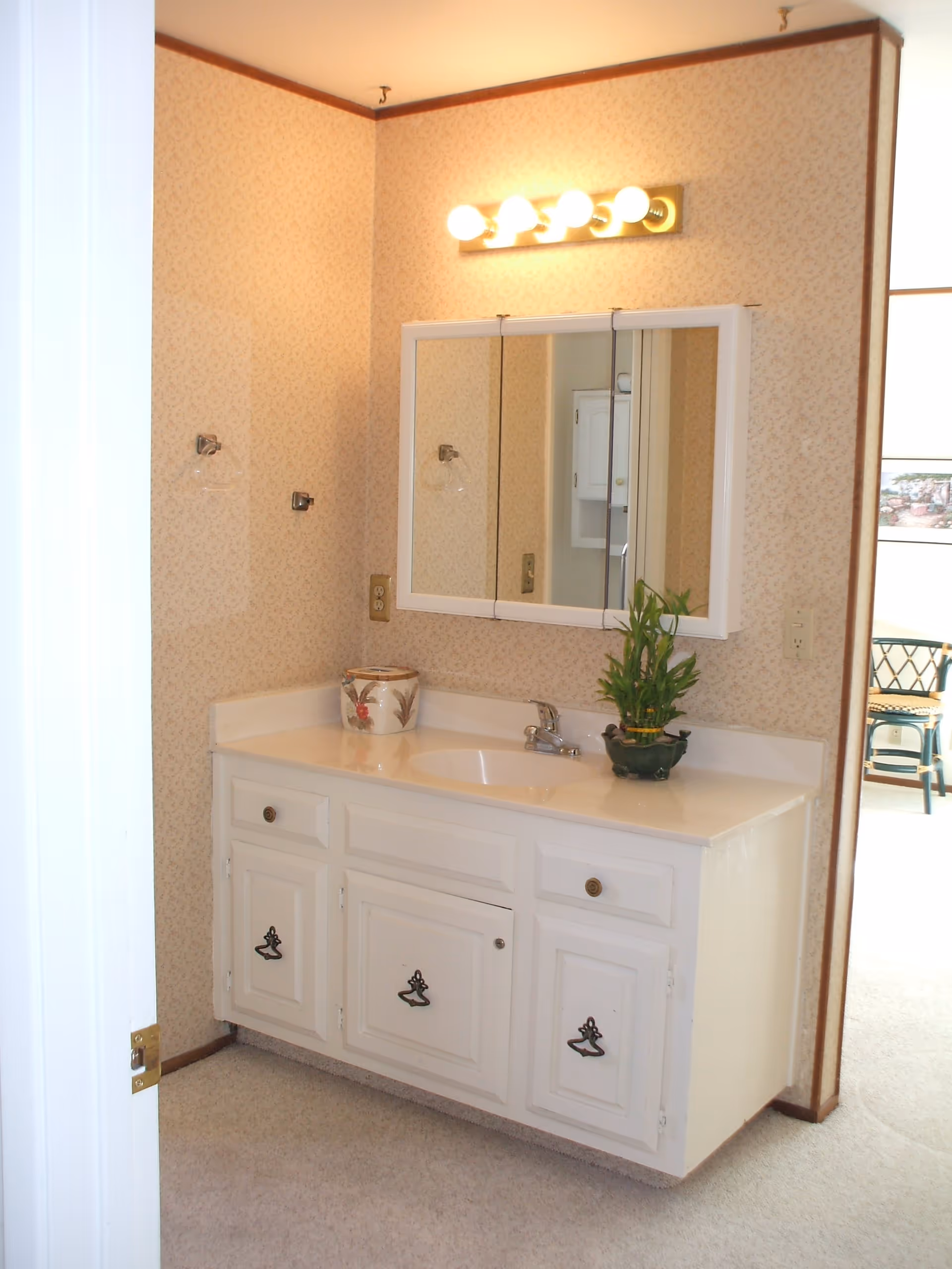 A bathroom vanity area with a white cabinet and countertop. There is a sink with a faucet, a decorative plant, and a ceramic container on the countertop. Above the vanity is a mirrored medicine cabinet with three panels and a light fixture with four bulbs. The walls have a light patterned wallpaper and the floor is carpeted. A partial view of an adjacent room with a chair is visible through the doorway.
