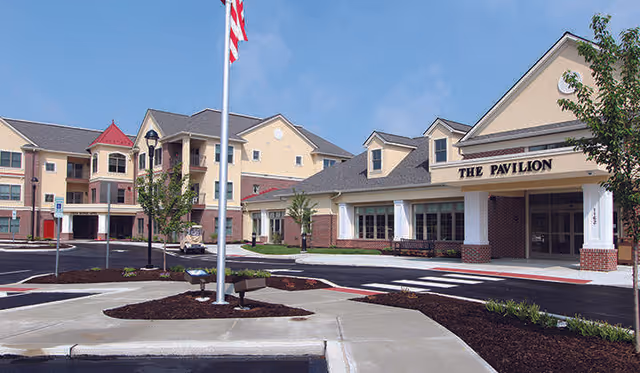Exterior view of Ware Presbyterian Village showing a multi-story residential building and a single-story building labeled 'The Pavilion' with an American flag on a flagpole in front, surrounded by landscaped areas and a parking lot.