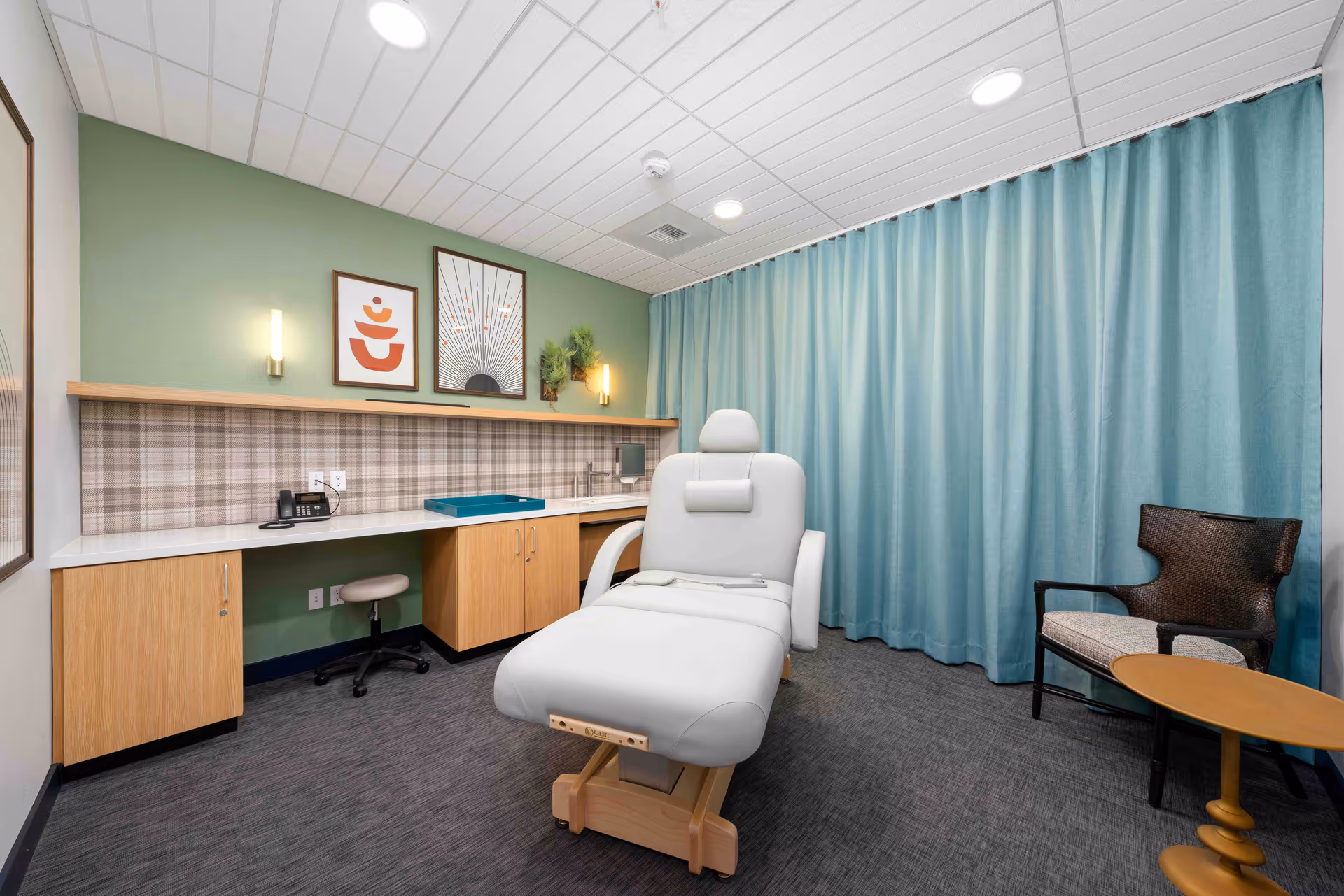 A treatment or therapy room with a white reclining chair in the center, a wooden desk with cabinets and a telephone against a green wall with framed artwork and wall sconces. A blue curtain covers one wall, and there is a woven chair with a cushion and a small wooden side table in the corner.