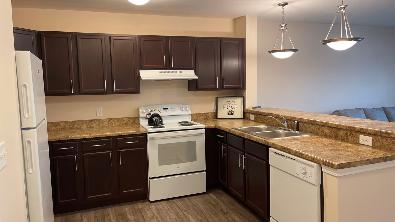 Open kitchen with dark wood cabinets, white appliances, a double sink and a breakfast bar overlooking a living area.