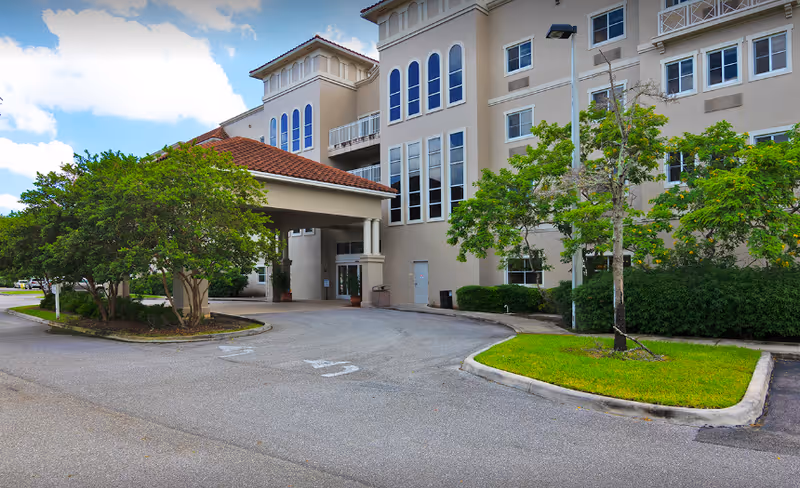 Exterior view of a multi-story senior living facility with beige walls and large windows. The building has a covered entrance with a red-tiled roof and is surrounded by green trees and landscaped grass areas. The sky is partly cloudy.